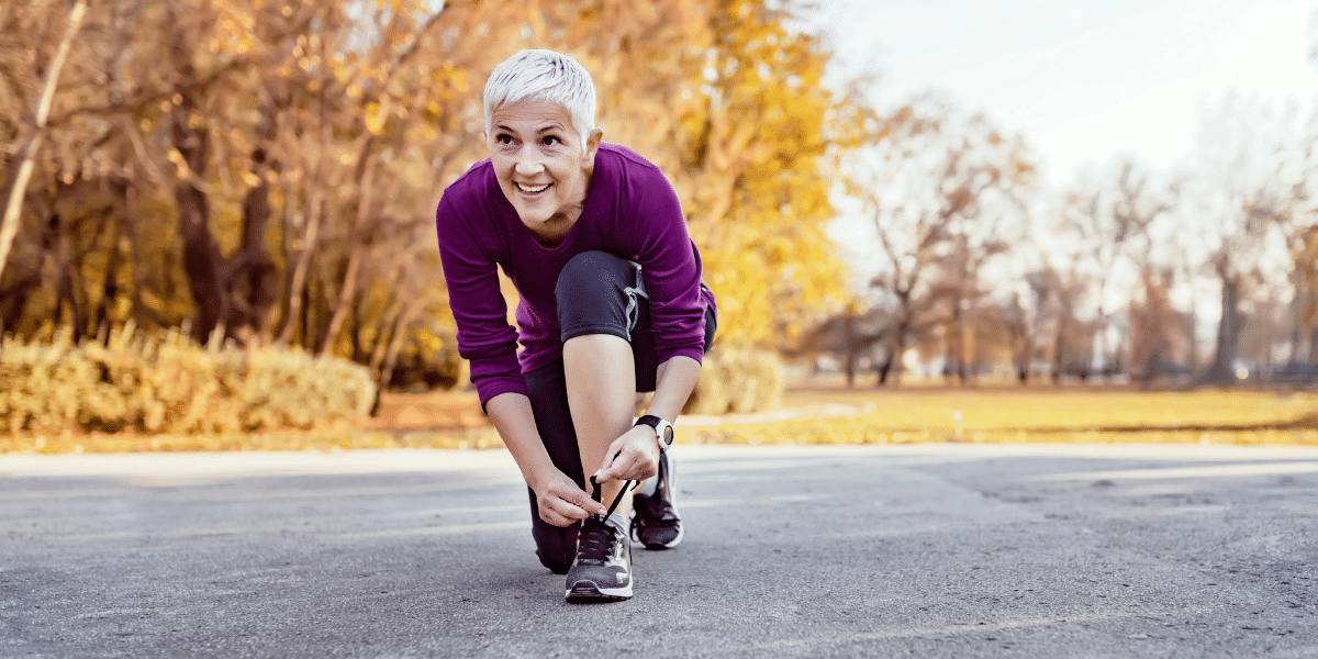 Older woman in purple top tying shoes in park on autumn day.
