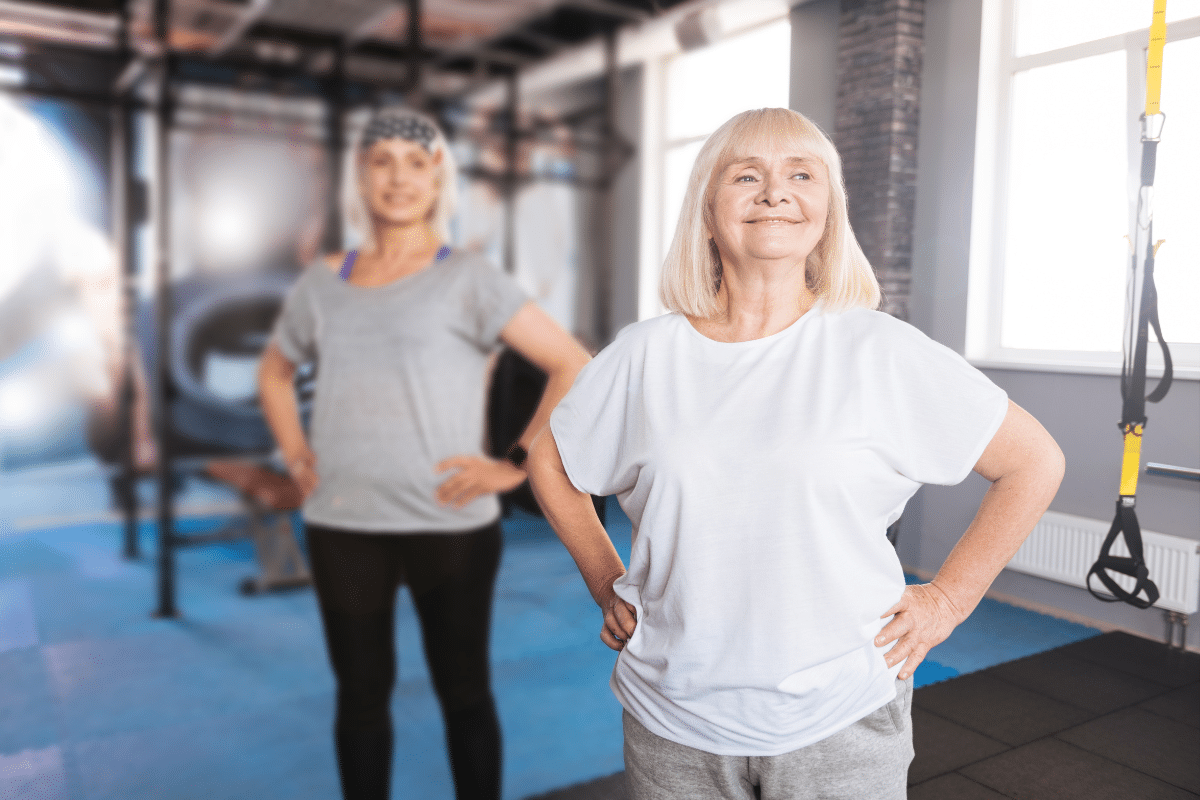 Two older women standing confidently with hands on hips in a gym setting, dressed in casual workout clothes and smiling, suggesting fitness and well-being.