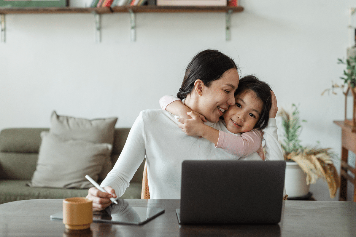 A mother showing love for her daughter while working on laptop.