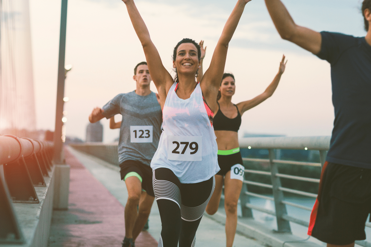 Group of runners crossing a finish line on a bridge at sunset, led by a smiling woman with race bib number 279 raising her arms in celebration.