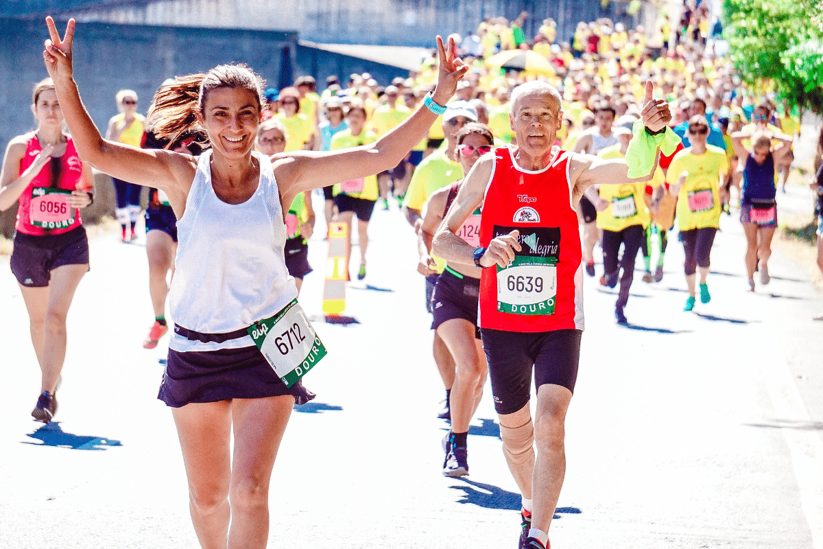Group of runners crossing a finish line on a bridge at sunset, led by a smiling woman with race bib number 6712 raising her arms in celebration.