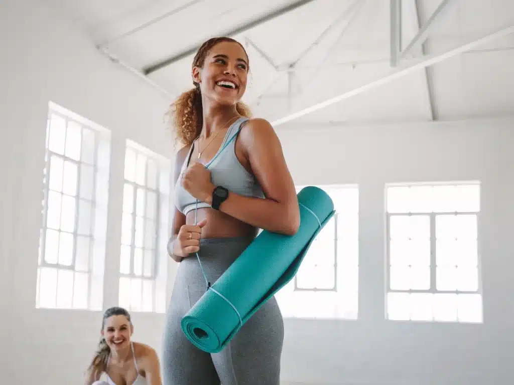 A women in yoga outfit, holding yoga mat and smiling towards someone.