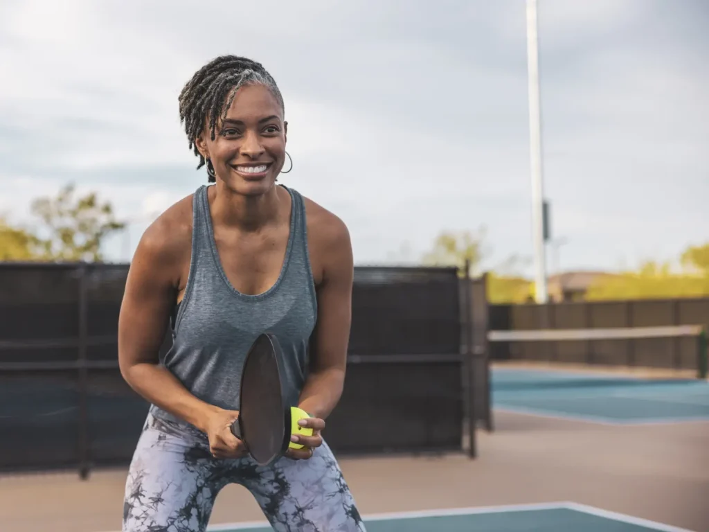 Woman smiling with pickleball paddle and ball outdoors.
